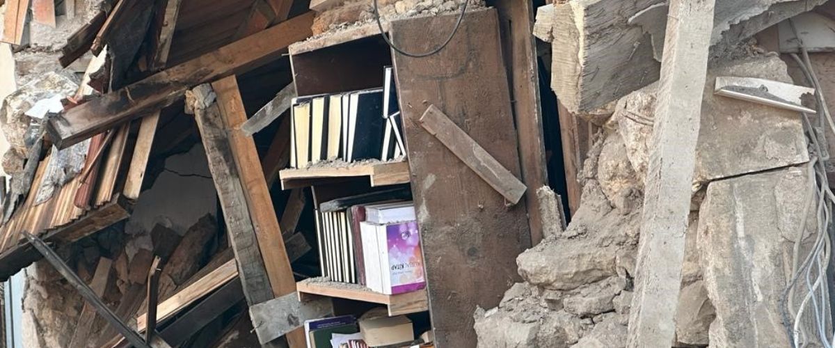 A bookshelf sits within the rubble of a collapsed building. 
