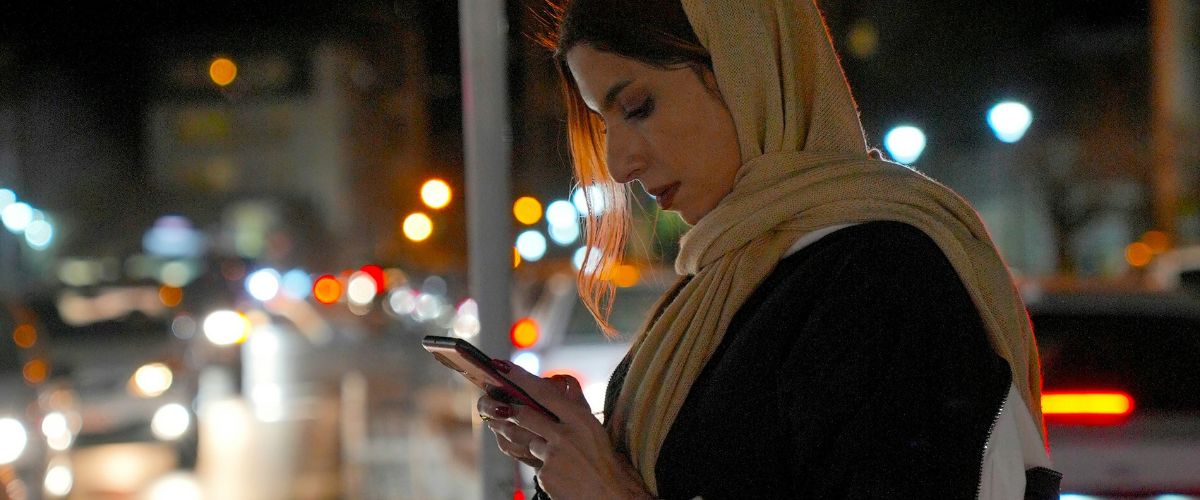 A women wearing a head covering looks down at her phone while in the city at night.