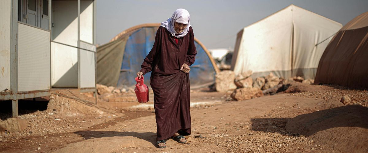 A woman in a refugee camp walks while lifting her skirt and holding a jug in her hand.